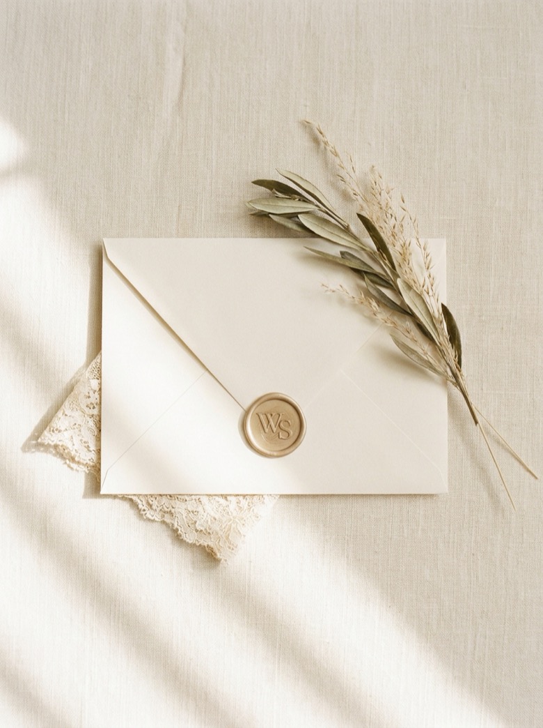 An ivory Wantwood Station wedding invitation sealed with a gold WS monogram wax seal, on cream linen with olive sprig and antique lace